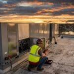 HVAC technician servicing a packaged rooftop unit on a commercial building at sunset